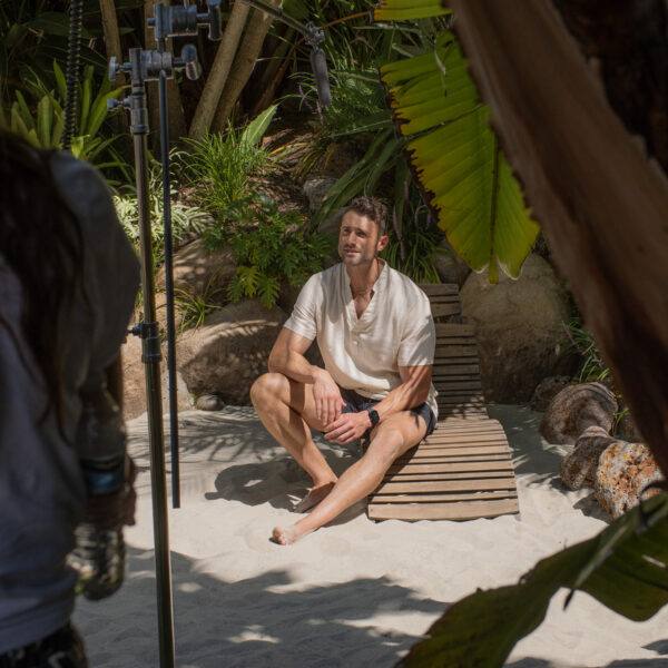 Talent sitting on lounger on White sand Beach with production crew in foreground at Tropic Shots production set
