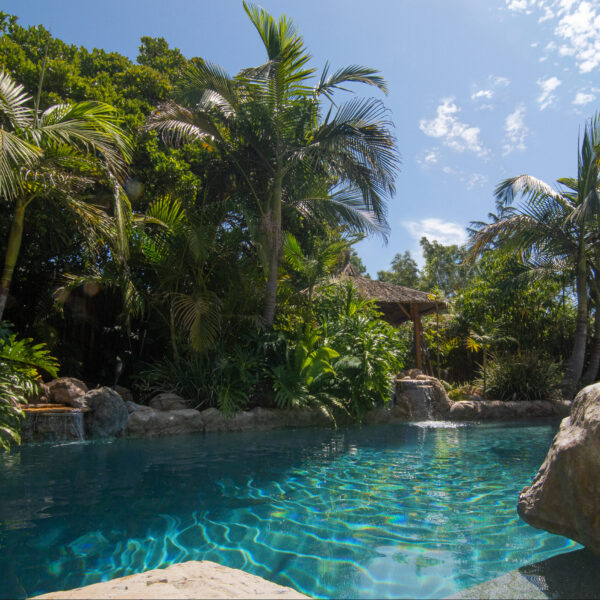 Crystal clear lagoon pool with dual waterfalls, surrounded by lush, tropical foliage at Tropic Shots production set