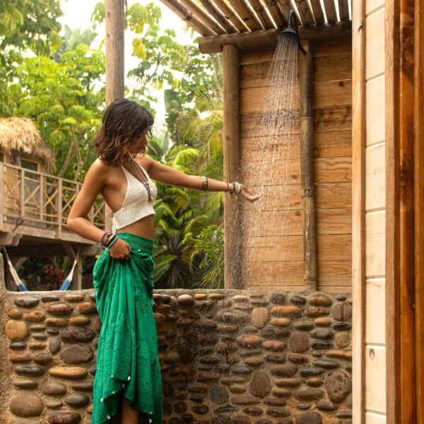 Model standing in front of outdoor cobblestone and Wood shower, checking water temp with treehouse and jungle trees in the background at Tropic Shots production set